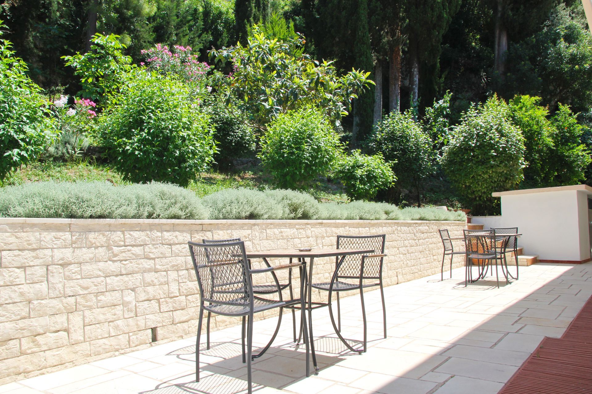 Αίθριο με μεταλλικά έπιπλα, πέτρινο τοίχο και πλούσια βλάστηση στο βάθος. Ηλιόλουστη μέρα.