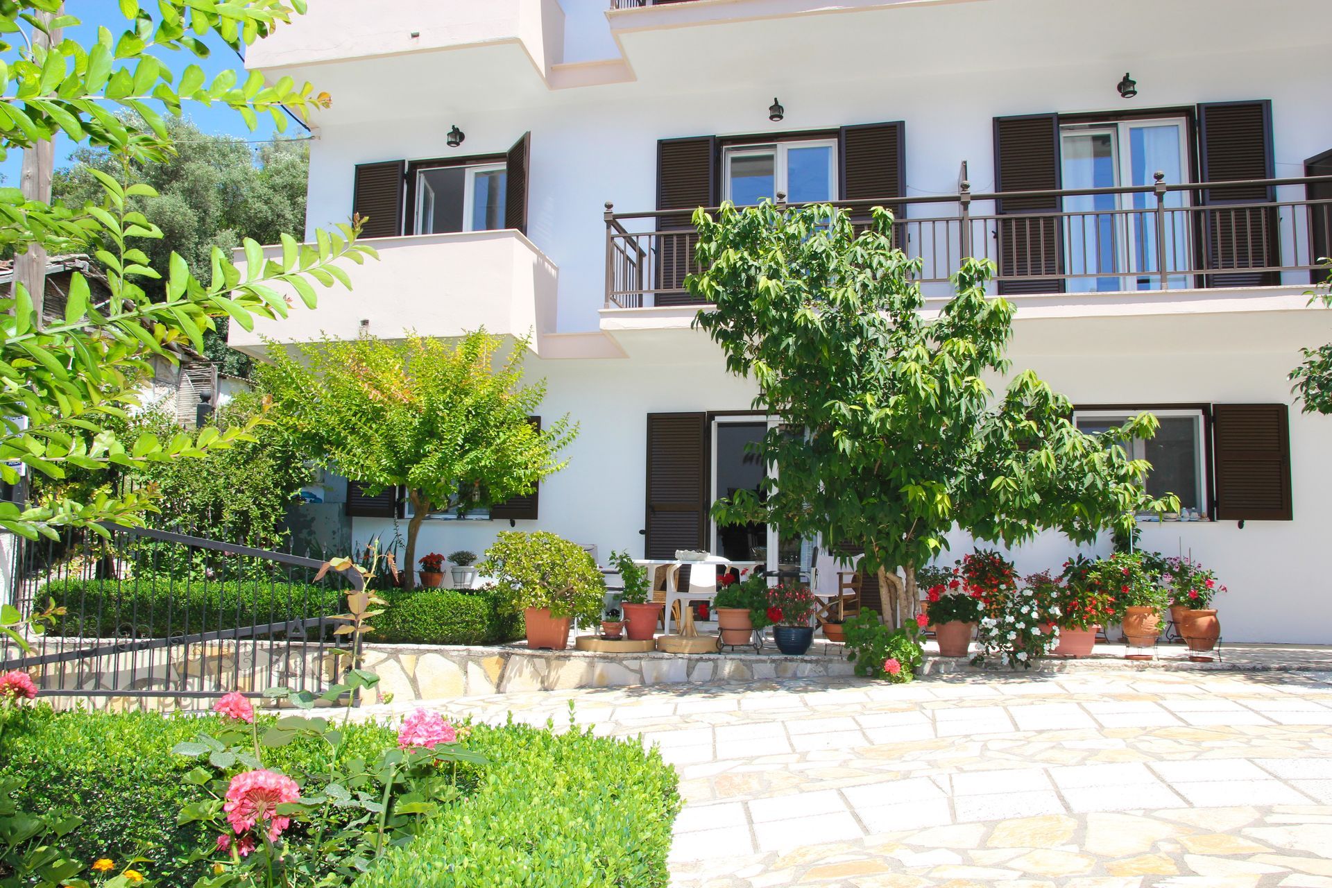 Λευκό κτίριο με καφέ παντζούρια και πέτρινη βεράντα, περιτριγυρισμένο από δέντρα και λουλούδια σε γλάστρες.