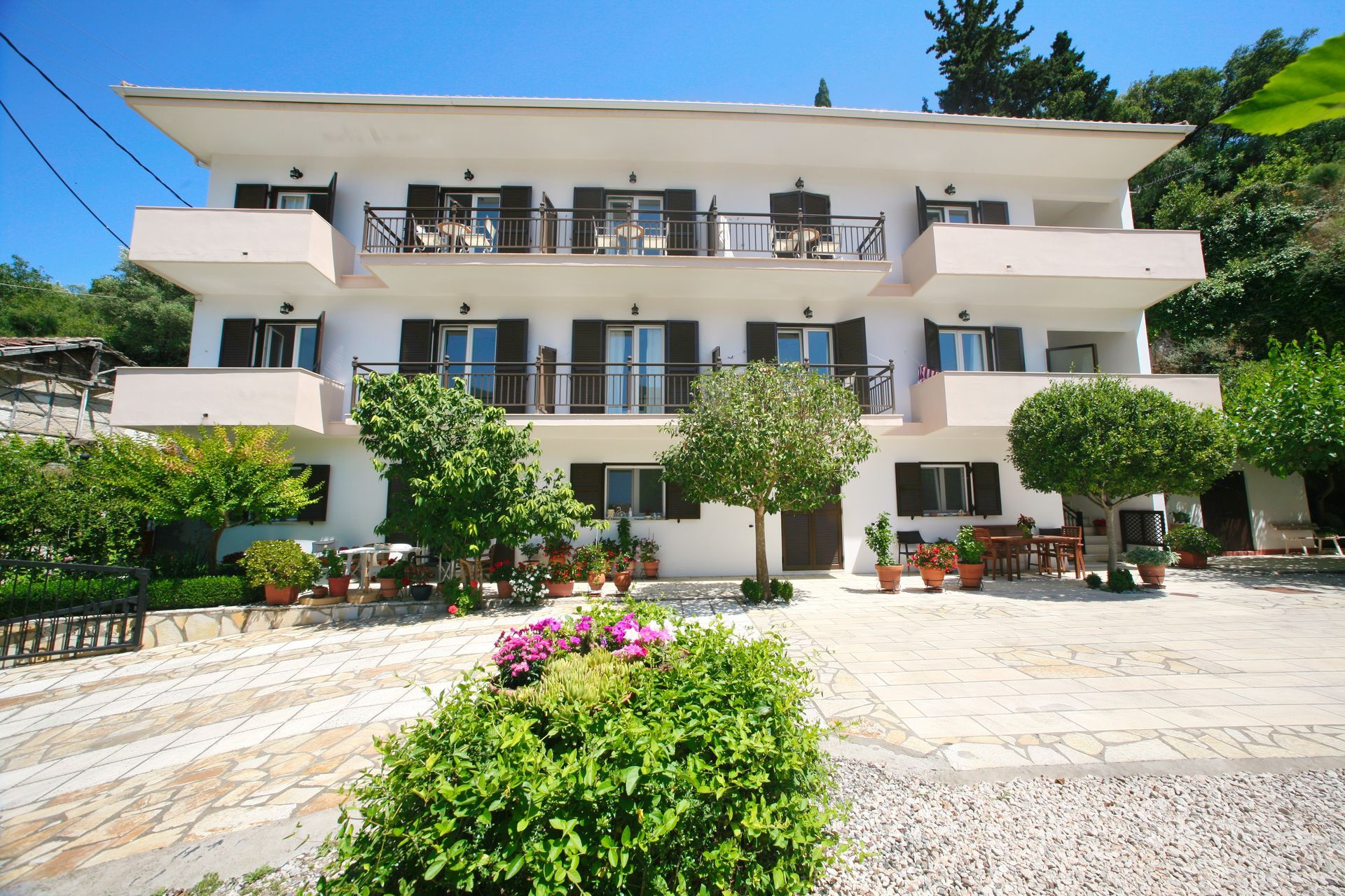 Λευκό πολυώροφο κτίριο με μπαλκόνια, περιτριγυρισμένο από πράσινο και πέτρινο δρόμο.