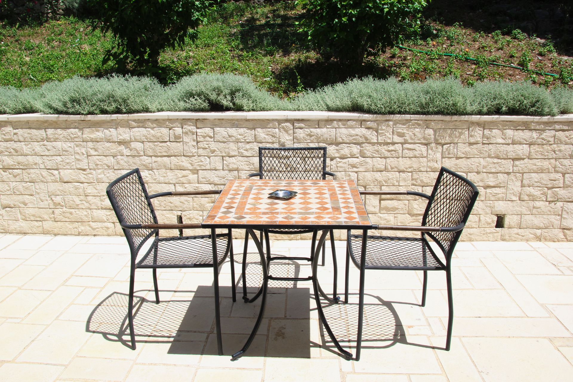 Τραπέζι και καρέκλες σε μια πέτρινη βεράντα, με έναν τοίχο και πρασινάδα στο βάθος.