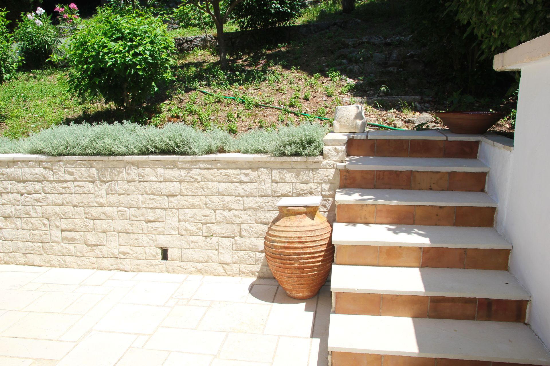 Πέτρινα σκαλοπάτια και τοίχος σε έναν ηλιόλουστο κήπο, με μια τεφροδόχο και πλούσια βλάστηση.