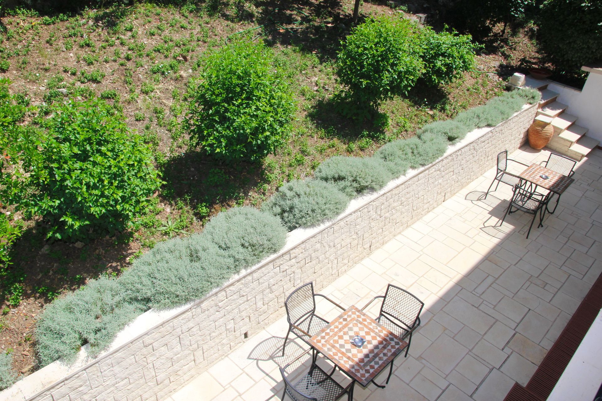 Αίθριο με τραπέζια και καρέκλες, δίπλα σε έναν κήπο με πράσινους θάμνους και χαμηλά, ασημένια φυτά.