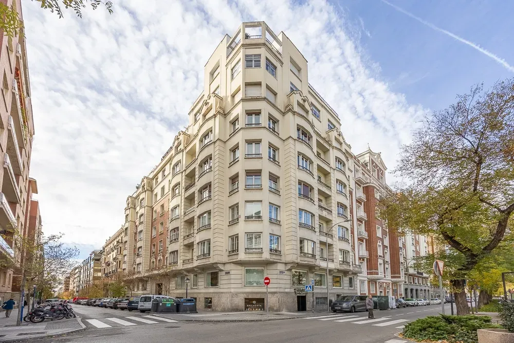 Un edificio de apartamentos de varias plantas, de color beige, situado en una esquina, con ventanas y balcones simétricos, en una intersección de calles de la ciudad.