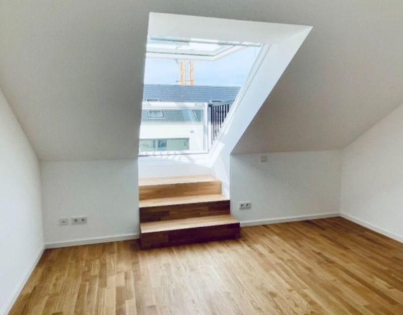 Helles Dachzimmer mit Holzboden und Dachfenster, über das eine kleine Treppe zu einer erhöhten Öffnung führt.