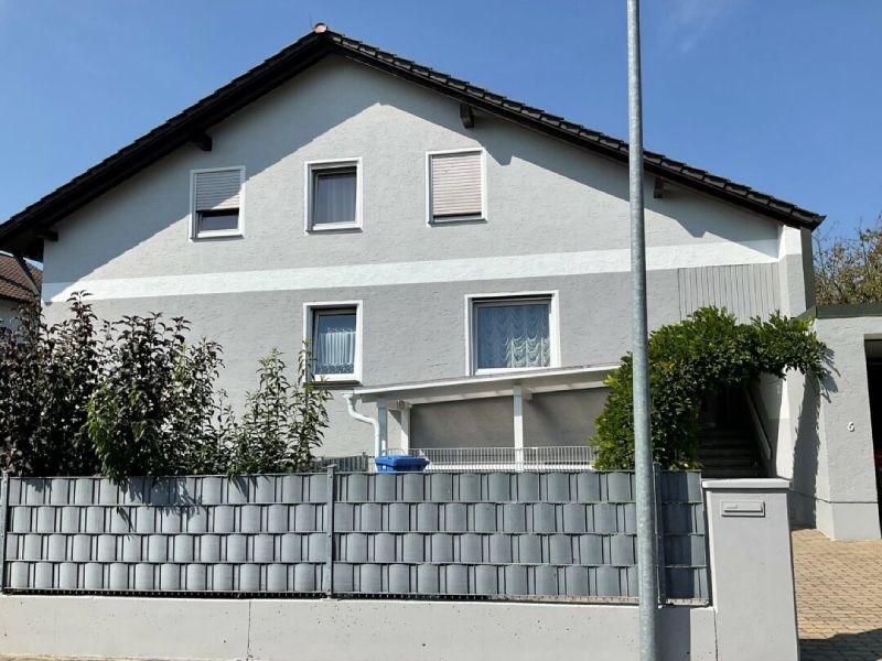 Zweistöckiges weißes Haus mit grauem Dach, Vorgartenzaun und kleinem überdachten Eingang in einer sonnigen Straßenszene