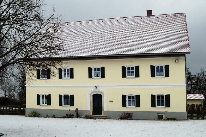 Schneebedecktes gelbes Haus mit grünen Fensterläden und dunkler Eingangstür in einer Winterlandschaft
