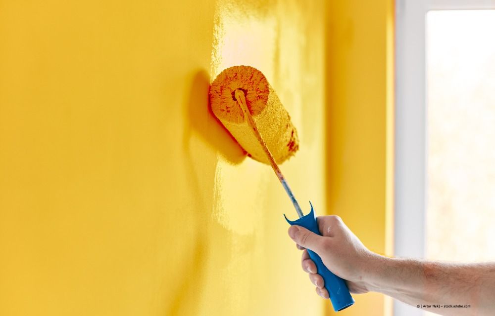 Eine Hand hält eine blaue Farbrolle und streicht eine leuchtend gelbe Wand.