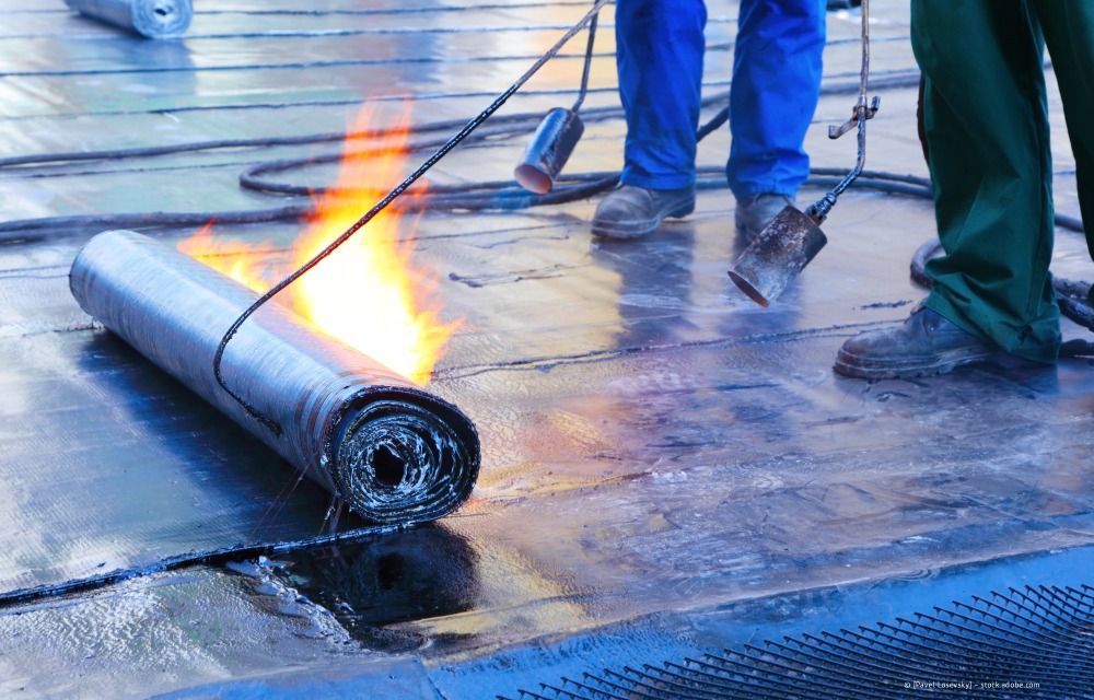 Arbeiter verlegen mit einem Schweißbrenner eine ausgerollte Dachmembran; Flammen sind auf dem nassen Dach sichtbar.