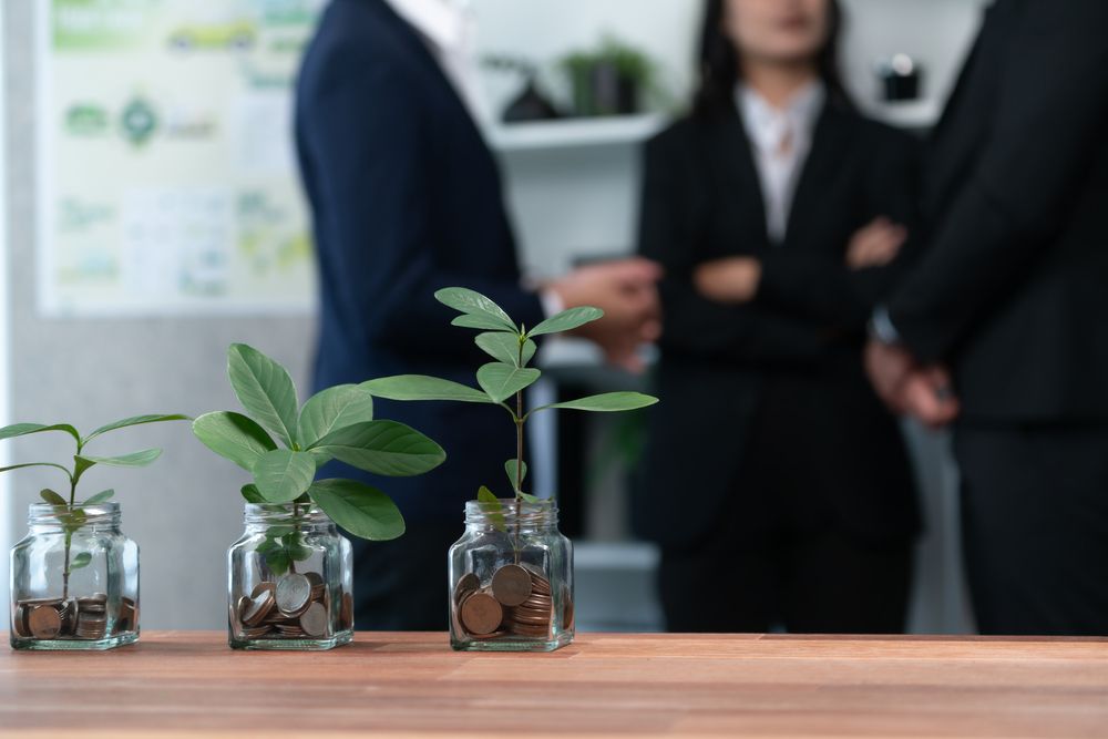 Un grupo de personas están de pie alrededor de una mesa con frascos llenos de monedas y plantas creciendo en ellos.