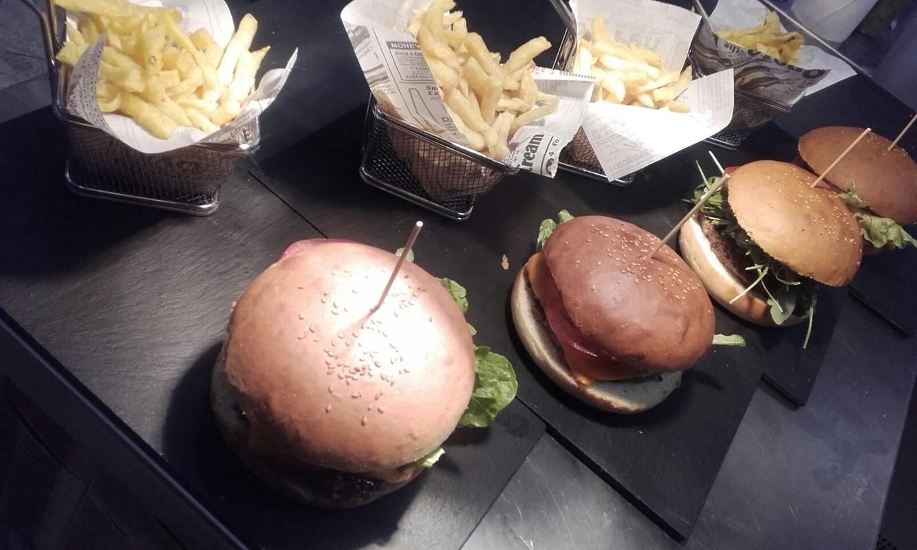 Tres hamburguesas con guarnición de patatas fritas servidas en pequeñas cestas metálicas sobre tablas individuales de pizarra negra.