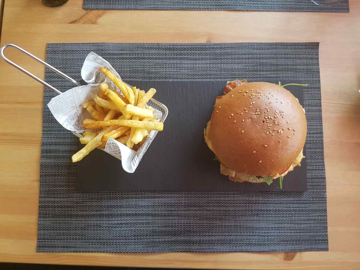 Una hamburguesa con pan de sésamo y una cesta de patatas fritas doradas sobre una tabla de piedra oscura, colocada sobre un mantel individual gris.