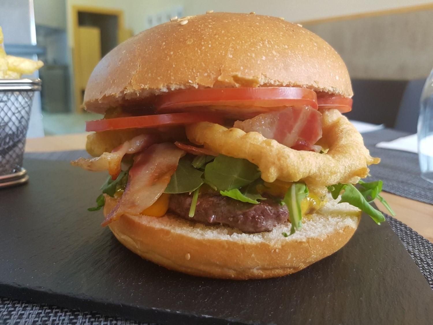 Una hamburguesa gourmet servida en una tabla de pizarra, cubierta con tocino, un aro de cebolla frita, rodajas de tomate y rúcula.