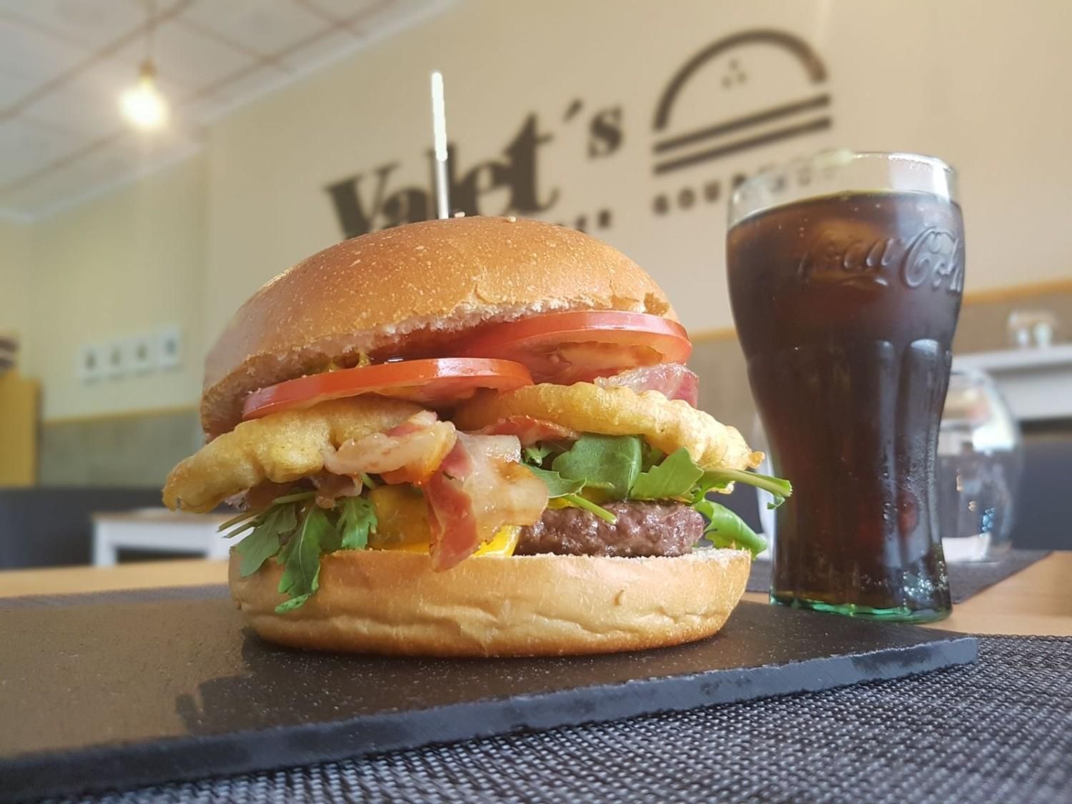 Una hamburguesa gourmet con aros de cebolla, tocino y verduras en una bandeja de pizarra oscura, servida con un vaso de Coca-Cola en un restaurante.