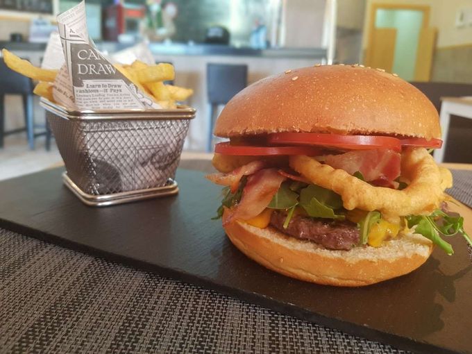 Una hamburguesa gourmet con queso, aro de cebolla, beicon y rúcula, acompañada de una pequeña cesta de patatas fritas sobre una pizarra.