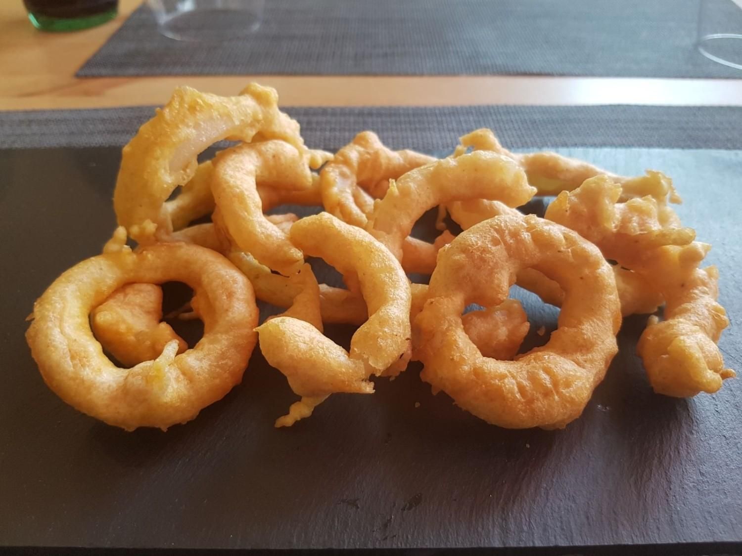 Un montón de aros de calamar fritos, de color marrón dorado, servidos en un plato de piedra oscuro y rectangular.
