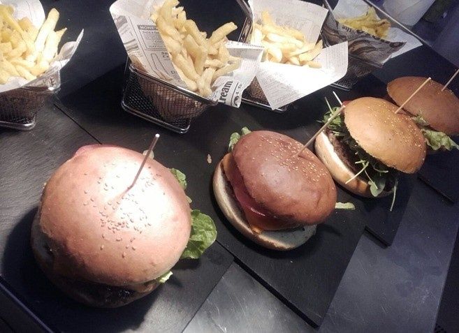 Tres hamburguesas y tres cestas de patatas fritas dispuestas en platos cuadrados negros sobre una superficie oscura.