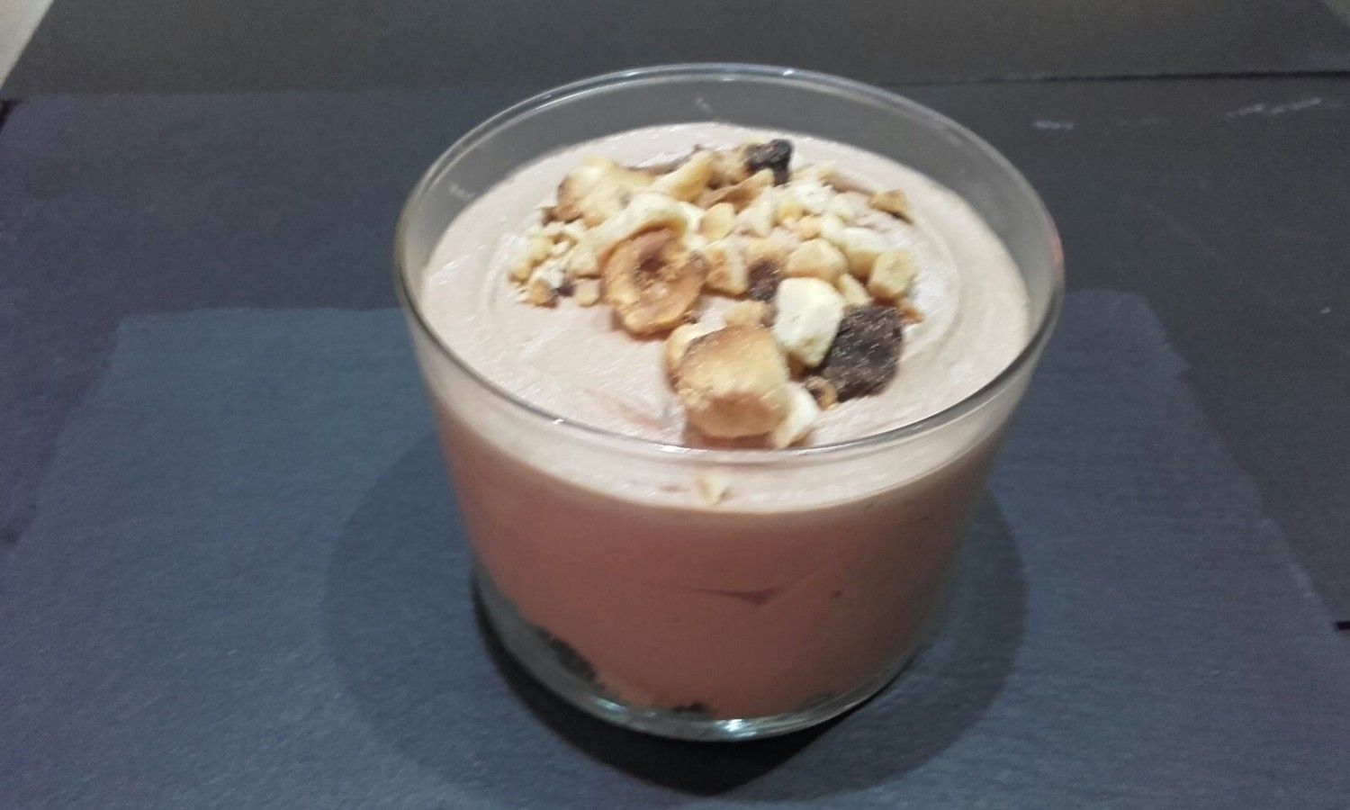 Una copa de cristal llena de mousse de chocolate cremoso, cubierta con avellanas picadas y trozos de chocolate sobre una superficie oscura.