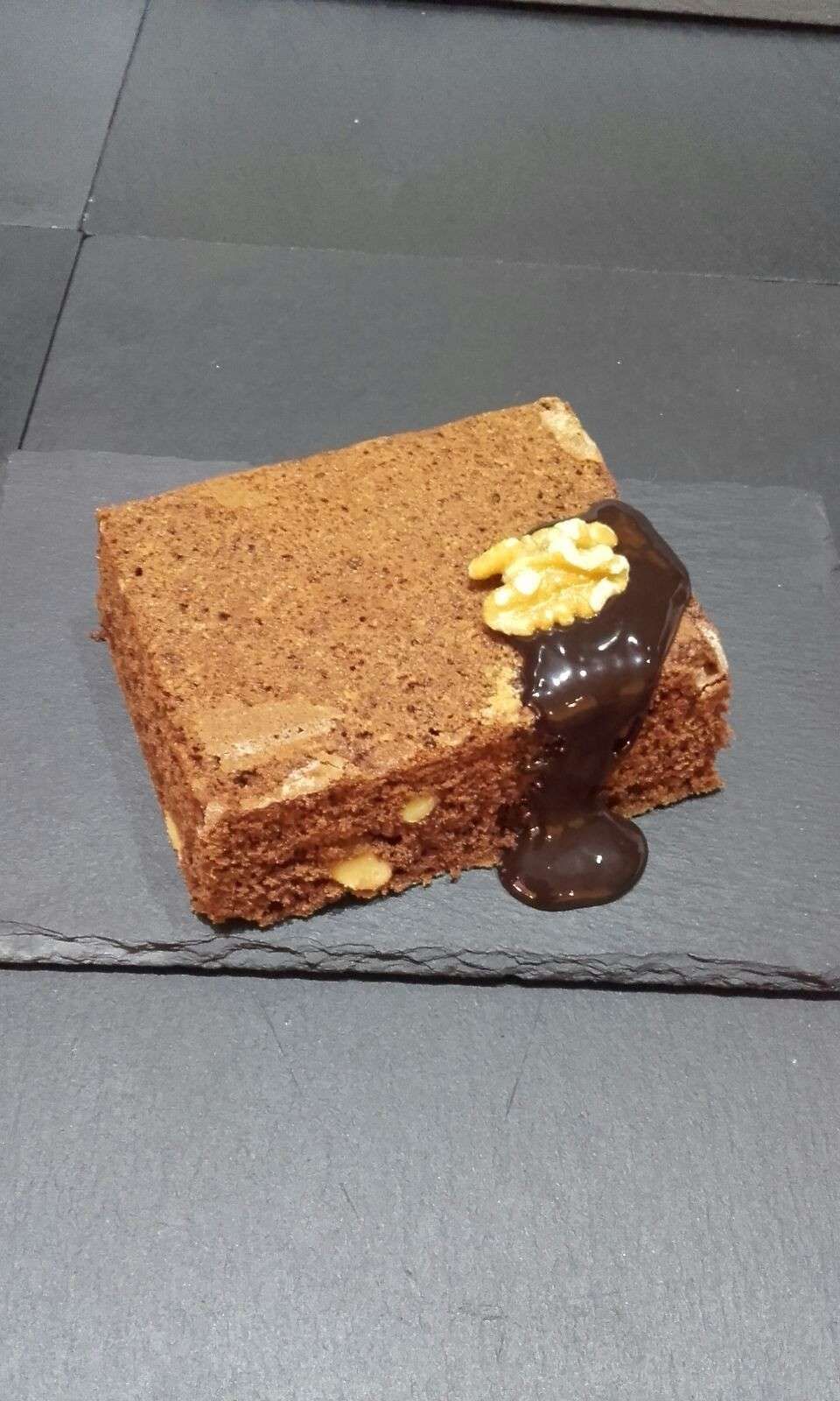 Un brownie cuadrado de chocolate cubierto con un chorrito de chocolate y una nuez, servido en un plato de pizarra gris.