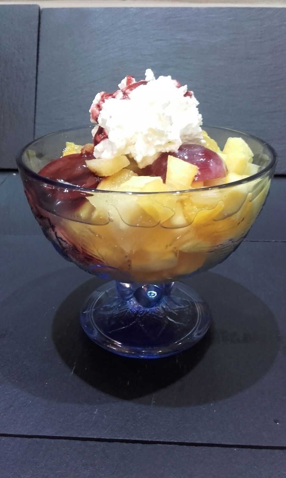 Un plato de postre de cristal lleno de piña amarilla cortada en cubos y cerezas de color rojo oscuro, coronado con una cucharada de crema batida blanca.