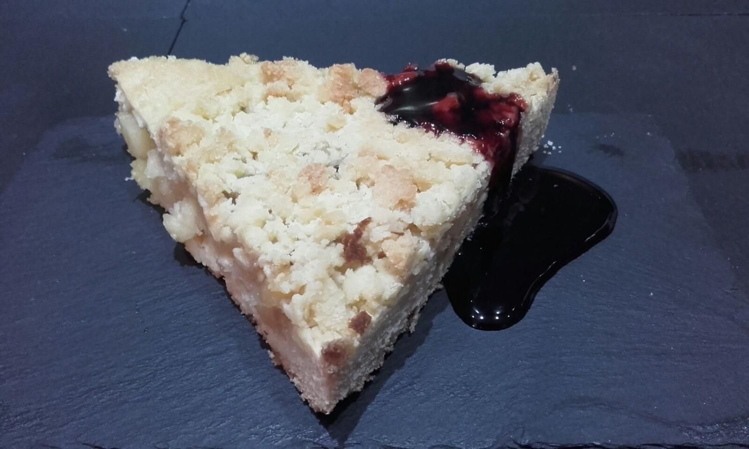 Una sola porción de pastel de frutas con crumble y salsa de frutos rojos oscuros, vertida sobre una superficie de pizarra negra.