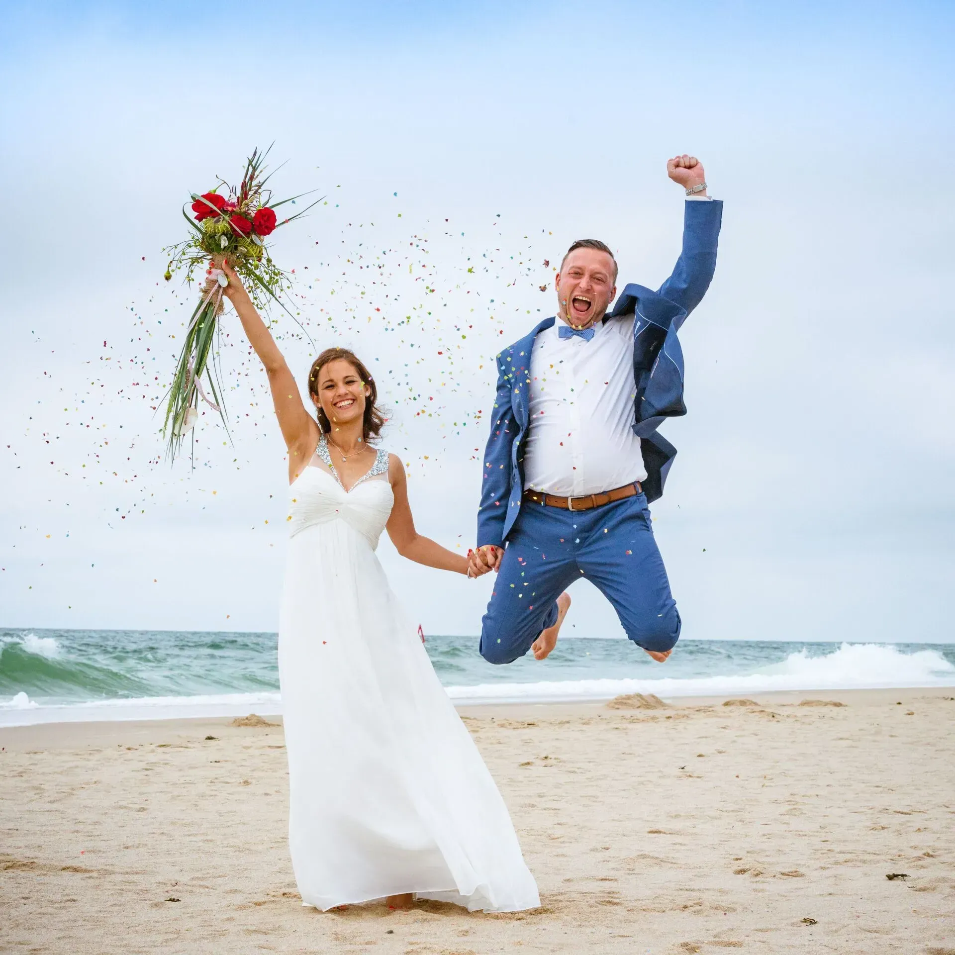 Ein Paar feiert am Strand eine Hochzeit; die Braut hält den Brautstrauß, der Bräutigam springt mit erhobener Faust.