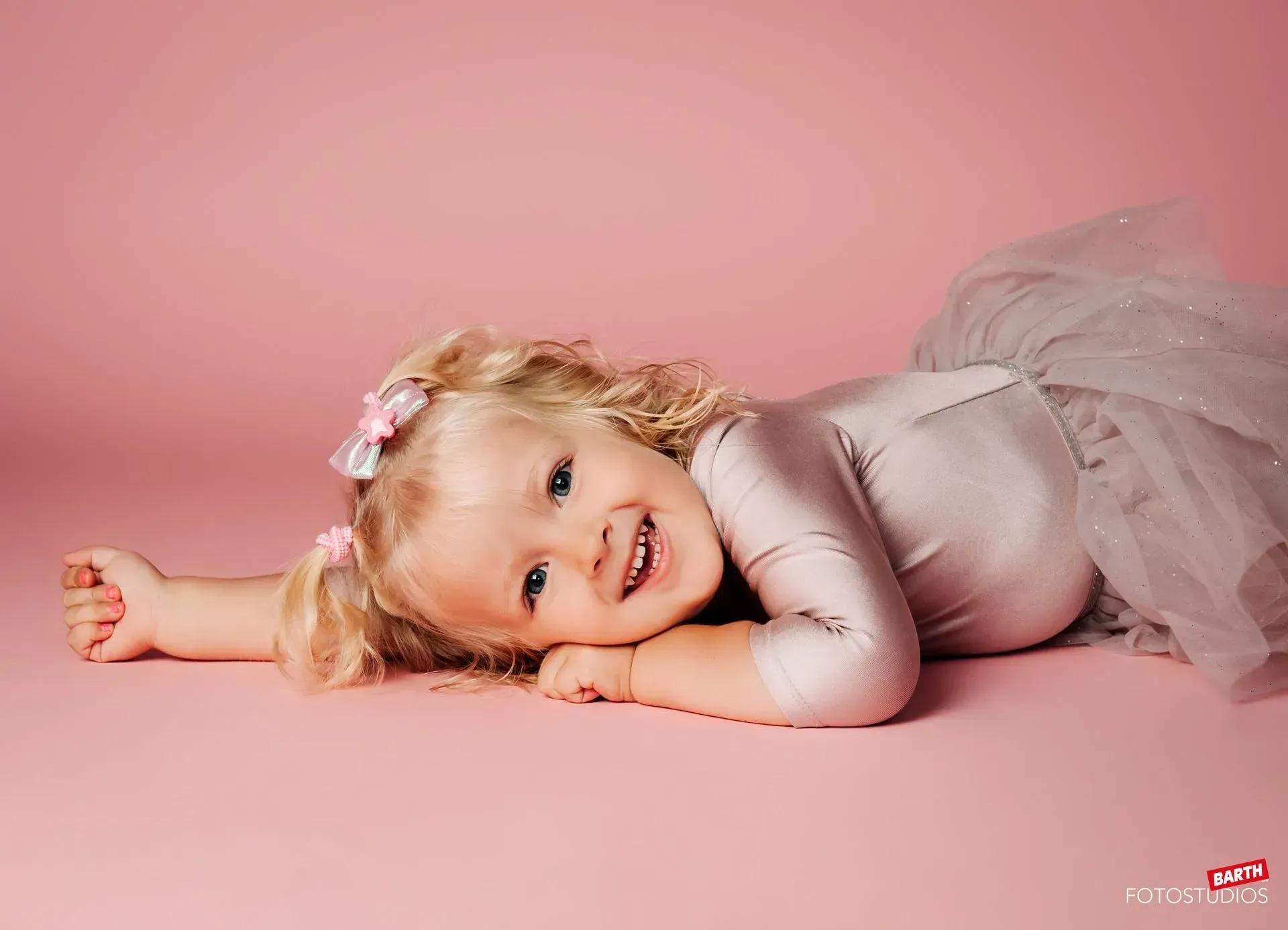 Ein lächelndes Kind in einem grauen Tutu-Kleid liegt auf rosafarbenem Hintergrund.