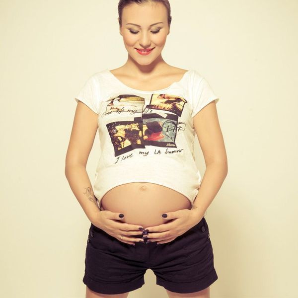 Lächelnde, schwangere Frau in weißem T-Shirt und schwarzen Shorts, die ihren Bauch hält, vor einem einfarbigen Hintergrund