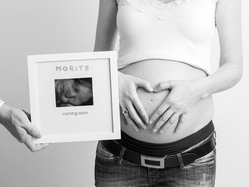 Schwangere Frau hält ihren Bauch neben einer Schachtel mit der Aufschrift „Moritz“ und einem Babyfoto.