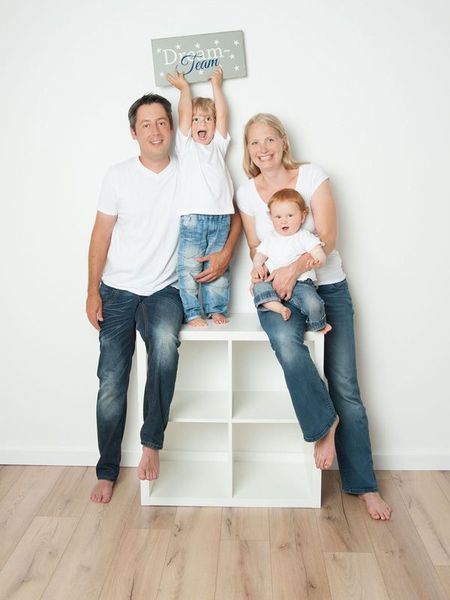 Eine Familie posiert mit einem Schild auf einem weißen Würfelregal in einem hellen Raum; zwei Erwachsene sitzen zusammen, ein Kind zwischen ihnen.