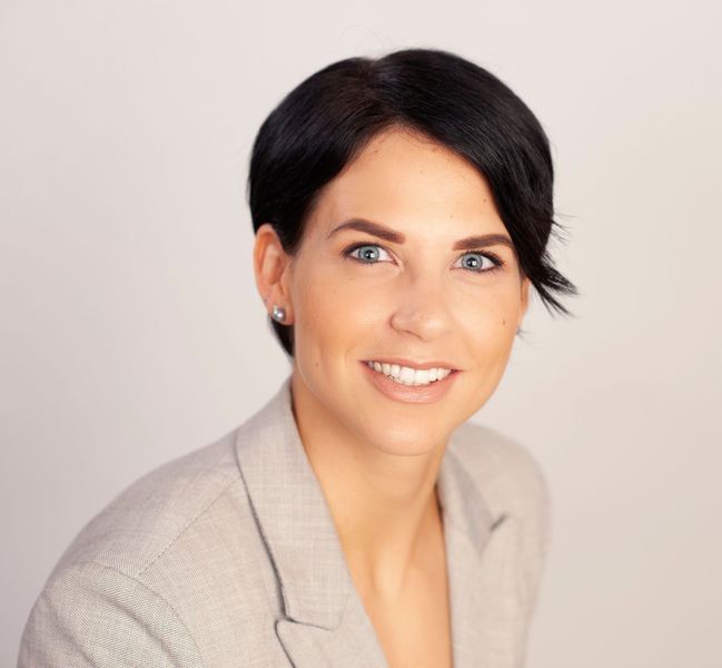 Lächelnde blonde Frau in einem schwarzen Blazer vor grauem Hintergrund