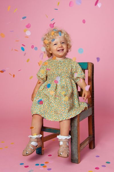 Ein lächelndes Kleinkind in einem geblümten Kleid sitzt auf einem Holzstuhl vor einem rosa Hintergrund, von dem bunte Konfetti herabrieseln.