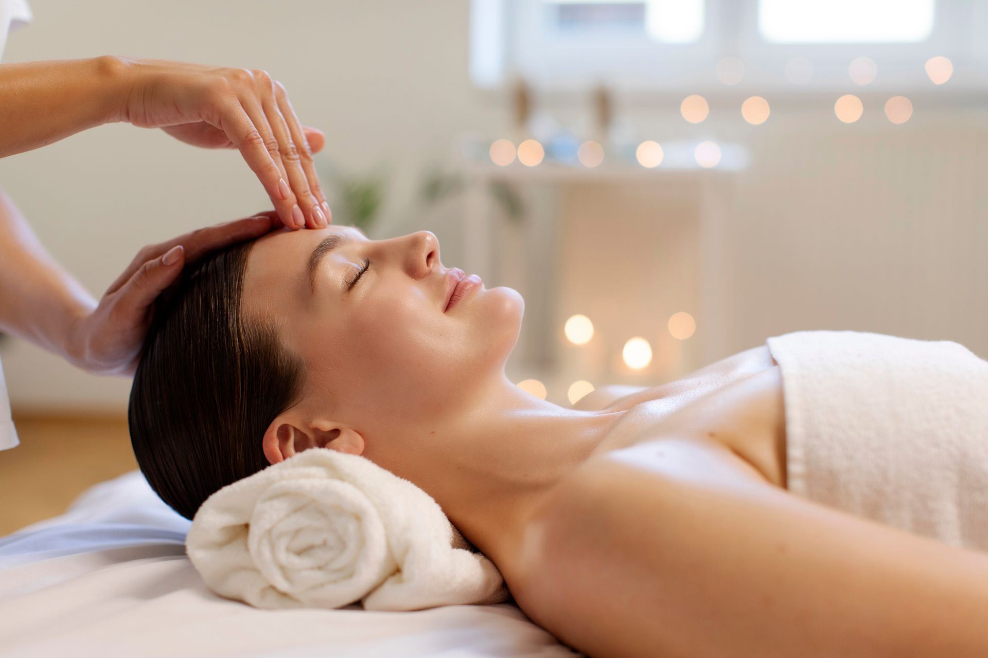 Femme recevant un massage du visage, les yeux fermés, serviette blanche, mains sur le front, ambiance spa.