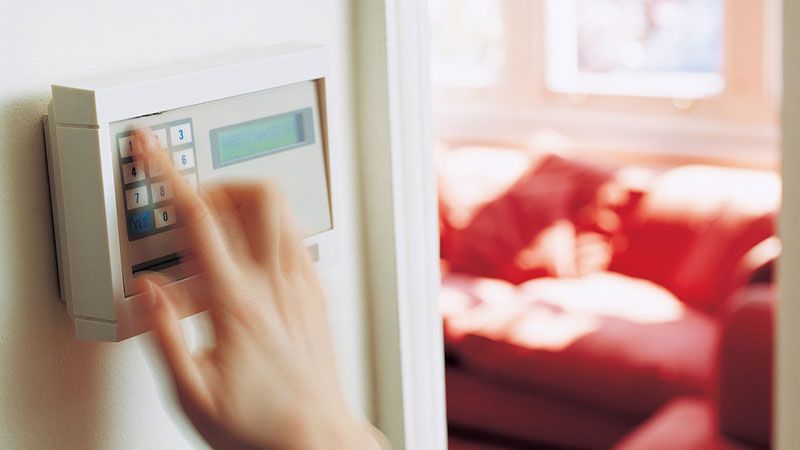 Une main atteint un thermostat mural, avec un canapé rouge ensoleillé en arrière-plan flou.
