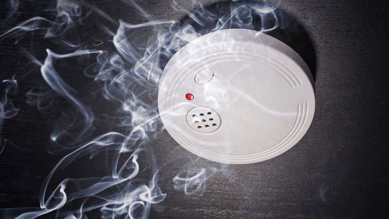 Détecteur de fumée blanc sur un plafond sombre, entouré de volutes de fumée.