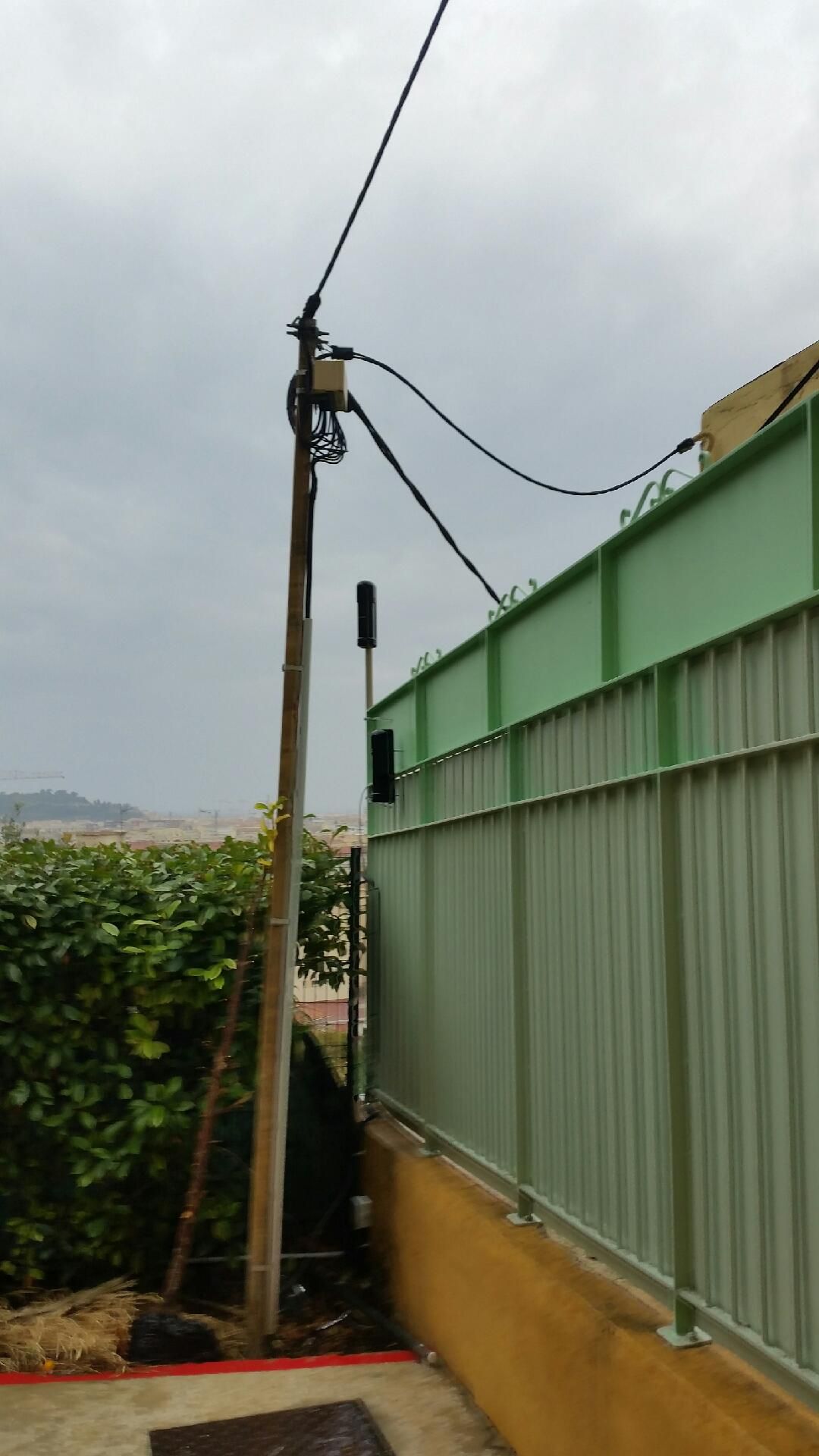 Étroite ruelle extérieure longeant une clôture verte et un poteau électrique sous un ciel nuageux.