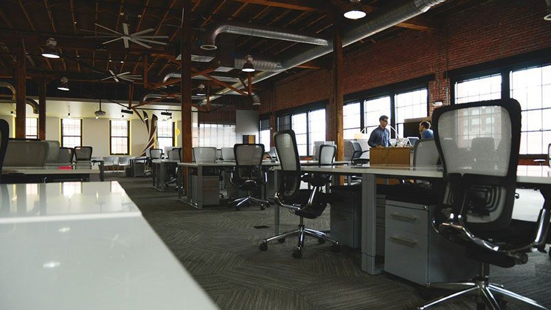 Espace de travail ouvert avec bureaux, chaises pivotantes et grandes fenêtres, en grande partie vide sous des éclairages industriels au plafond.