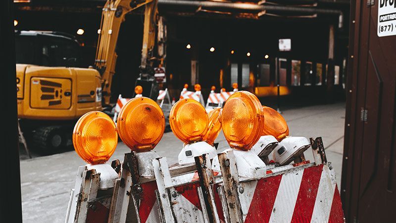 Des feux de chantier orange éclairent des barrières rayées la nuit, avec une pelleteuse jaune en arrière-plan.