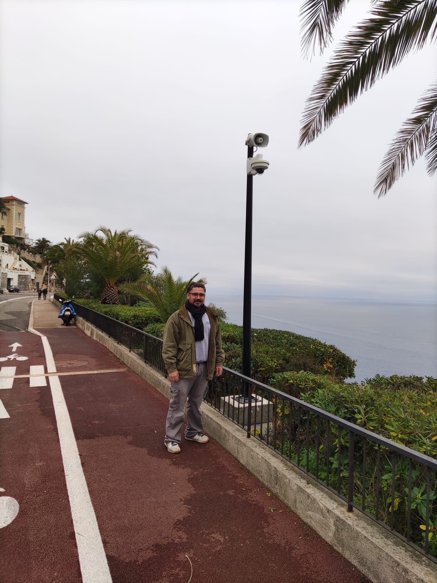 Une personne se tient sur une promenade en bord de mer, à côté d'une rambarde et d'un lampadaire, avec vue sur l'océan et des palmes.