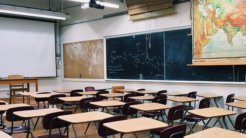 Salle de classe vide avec des rangées de pupitres, un tableau noir, un écran de projection déroulant et une carte du monde au mur.