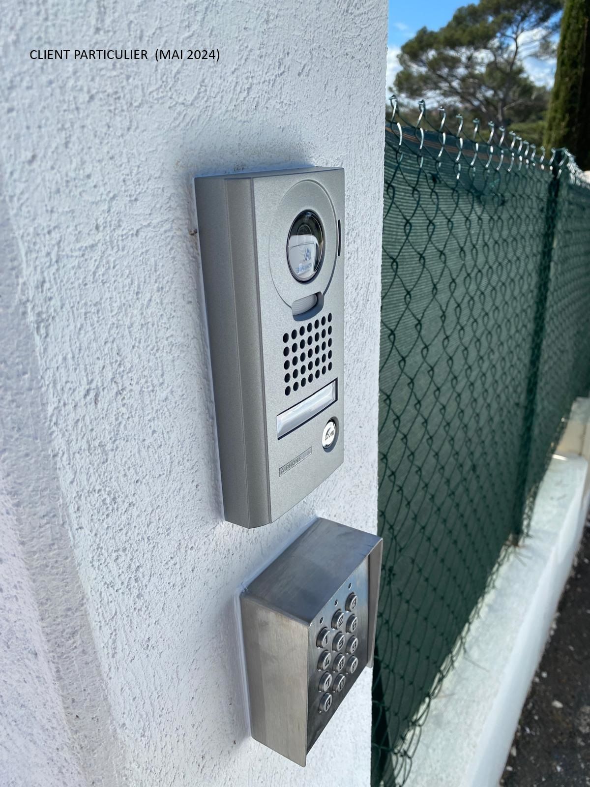 Interphone mural et boîtier d'appel métallique à côté d'un mur en stuc blanc et d'une clôture en grillage.