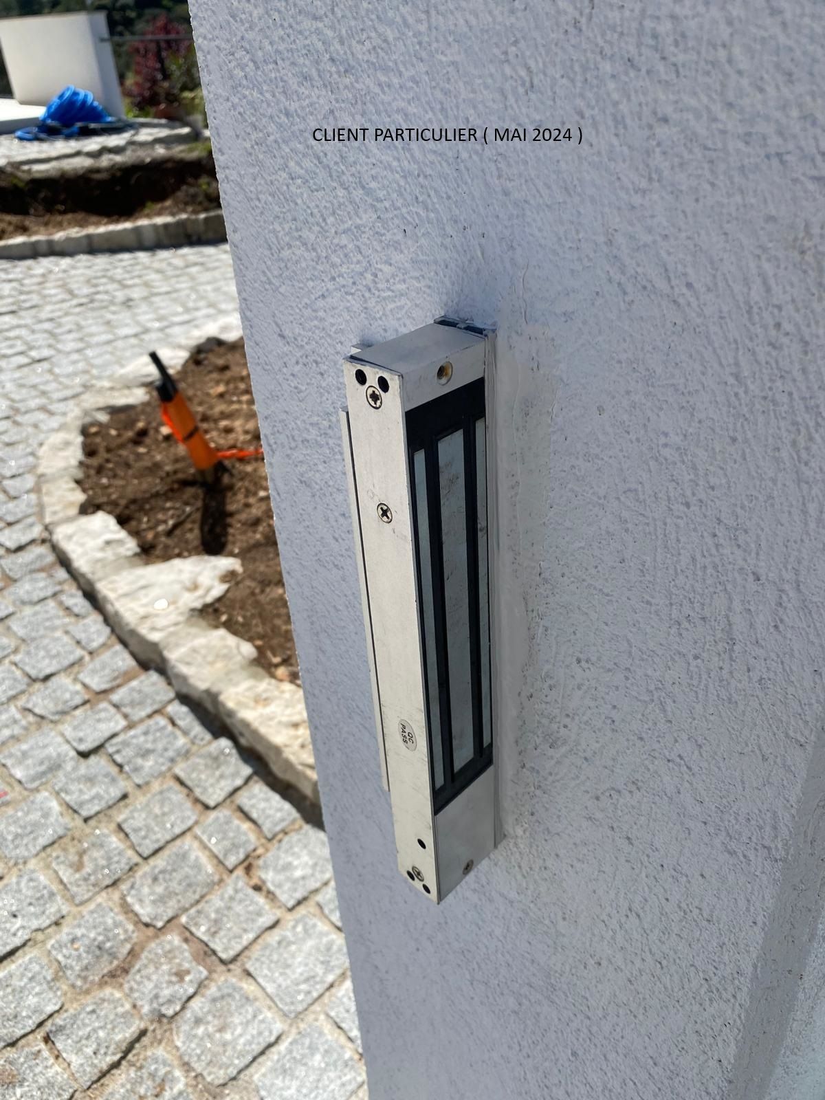 Boîte aux lettres ou boîte à colis métallique fixée au mur, sur un mur en stuc blanc, à côté d'un chemin pavé.