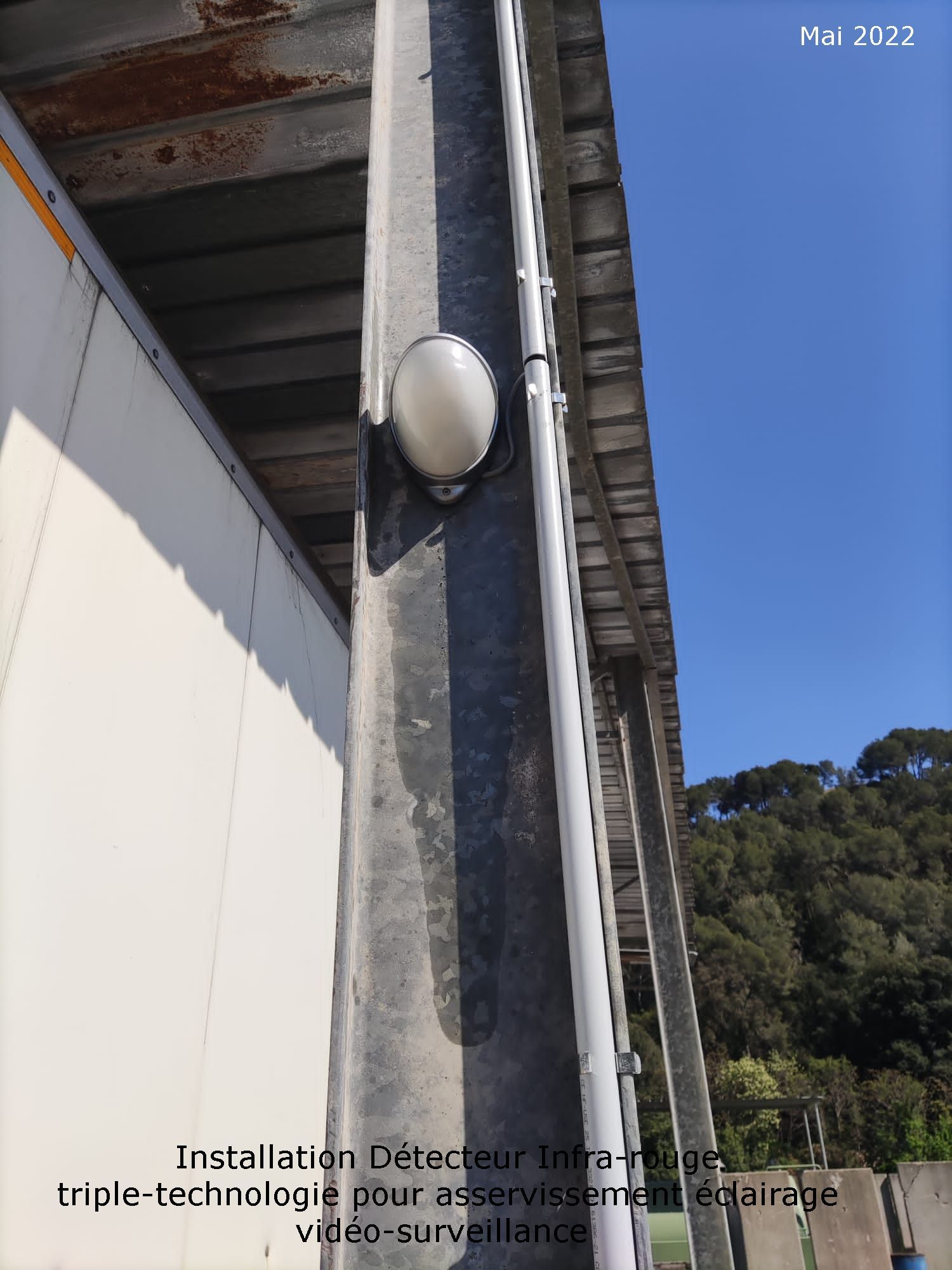 Vue extérieure d'un angle de bâtiment avec un mur blanc, une colonne en béton et un luminaire rond encastré sur fond de ciel bleu