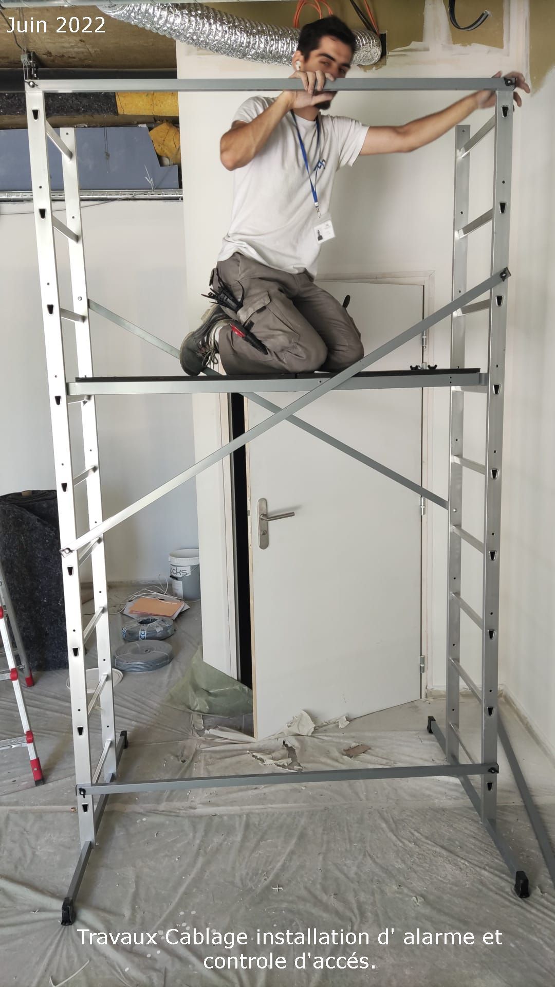 Un ouvrier, agenouillé sur un échafaudage métallique à l'intérieur d'une pièce en rénovation, perce le plafond.