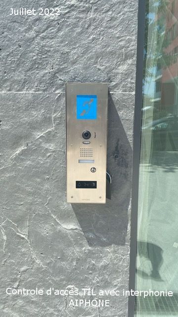 Interphone mural avec écran bleu et boutons, placé à côté d'une porte vitrée sur un mur gris texturé.