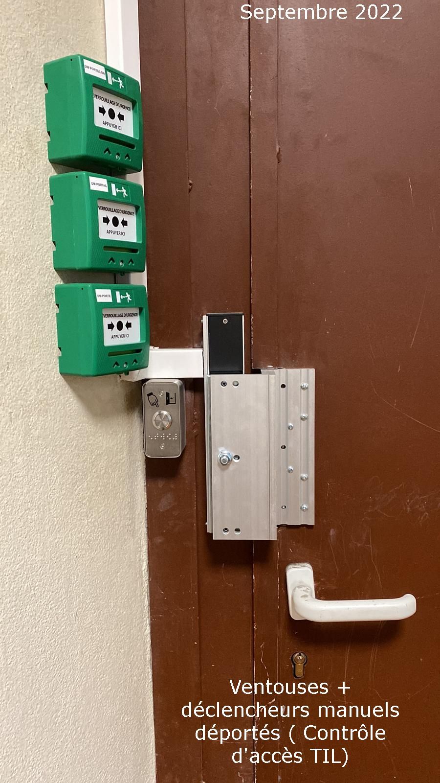 Porte marron avec interphone, clavier numérique et boîtes aux lettres vertes superposées contre un mur et une rampe.