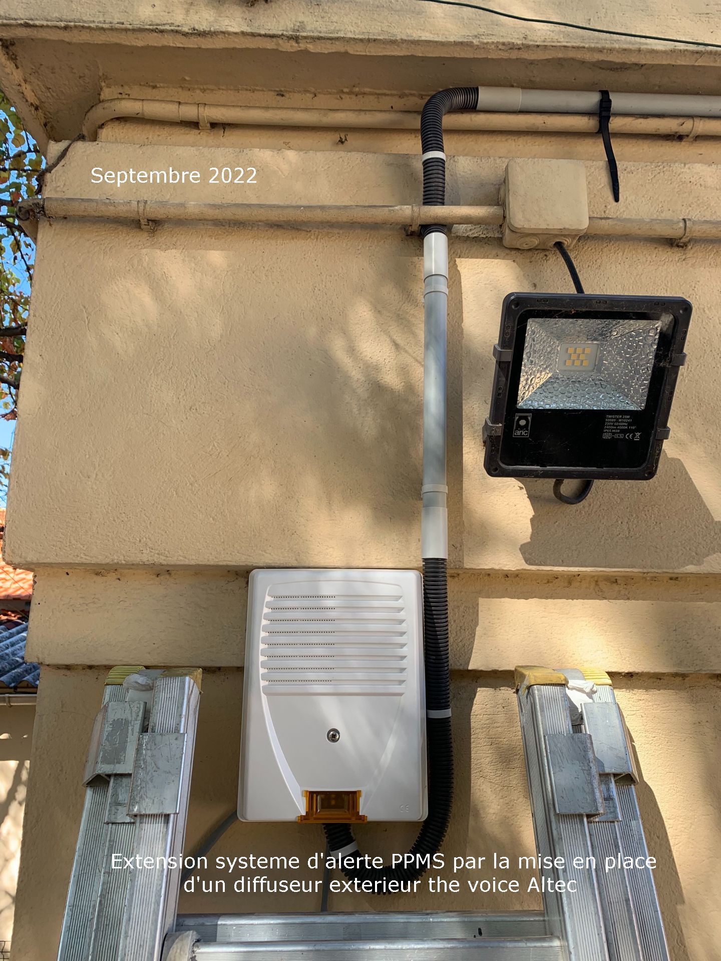 Mur extérieur avec compteur électrique, conduits de ventilation et projecteur noir sur un bâtiment beige.