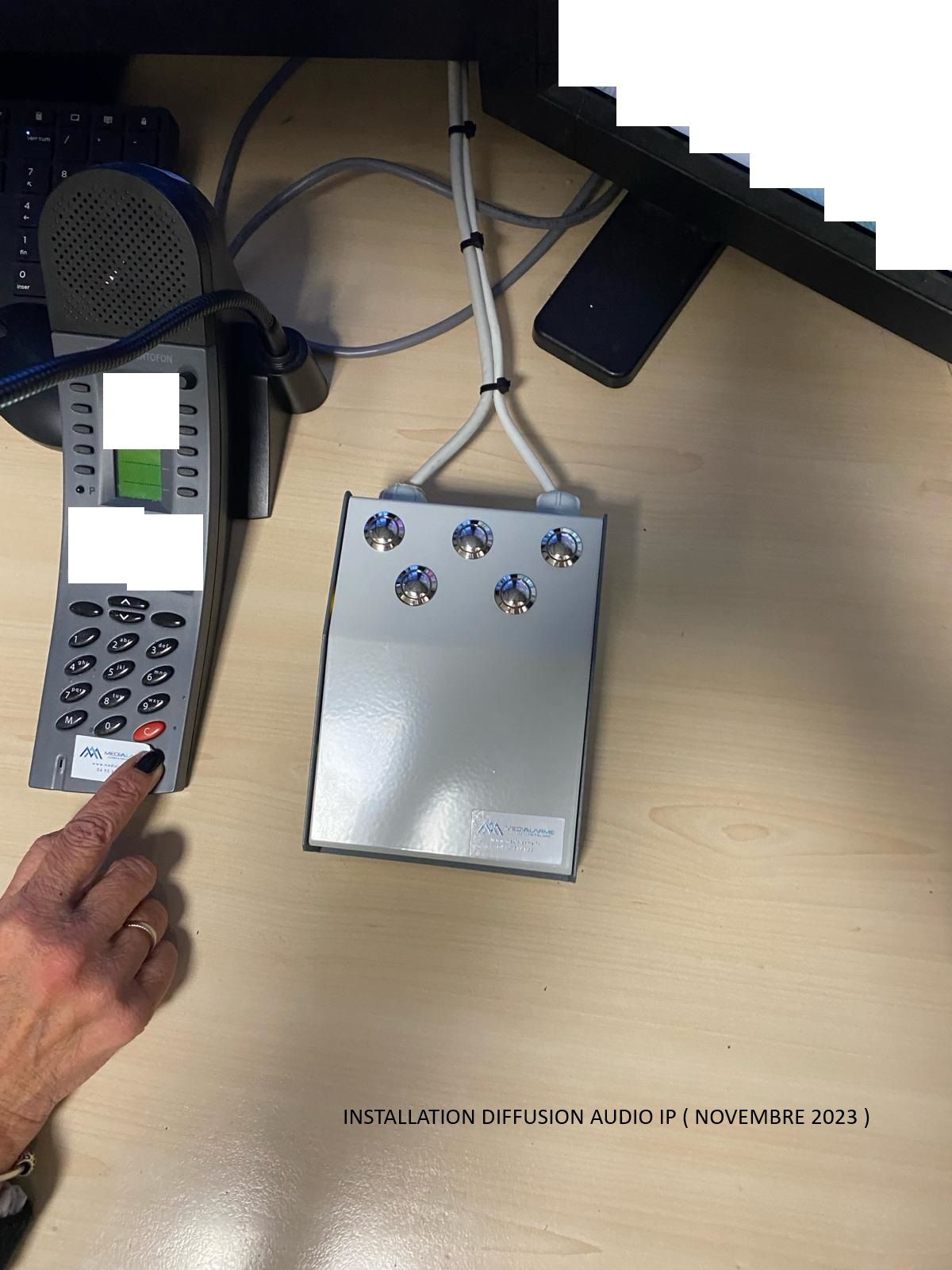 Main appuyant sur le clavier d'un téléphone à côté d'un petit boîtier de commande métallique avec des boutons bleus et des câbles connectés
