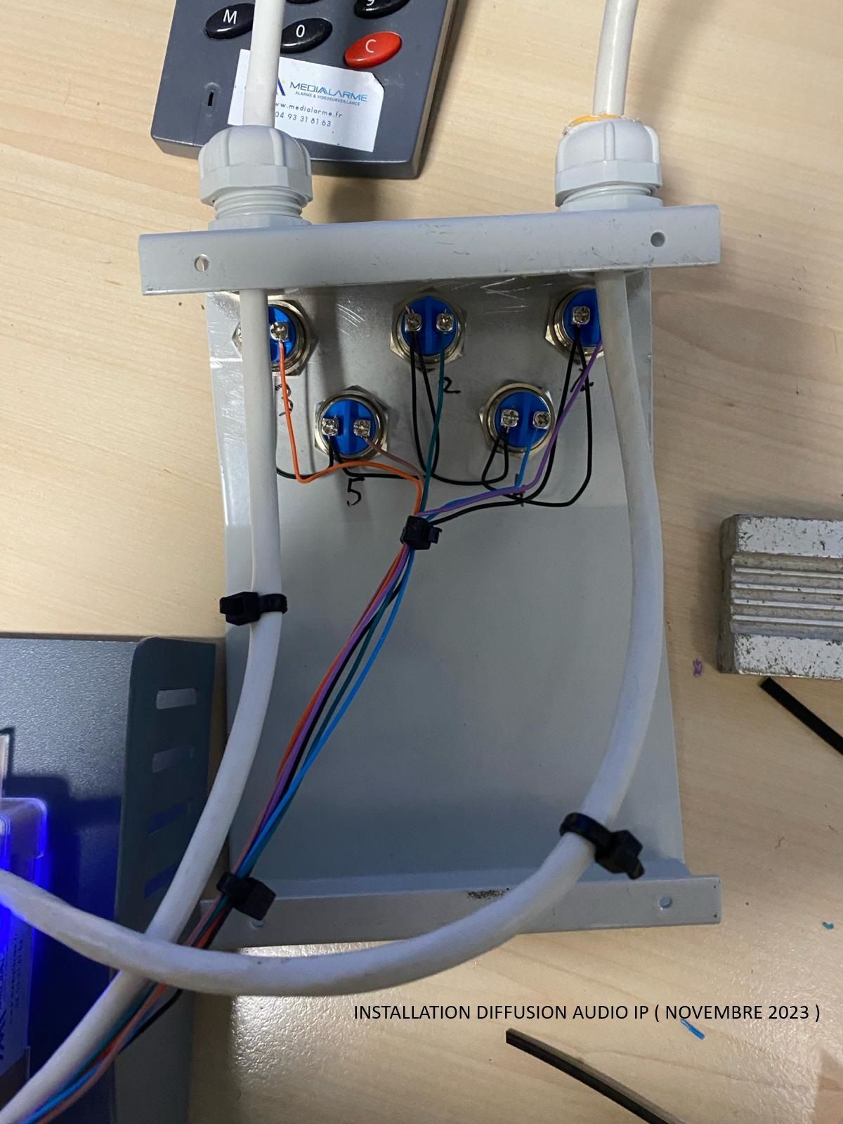 Boîte de jonction électrique ouverte avec câblage bleu et tuyaux raccordés, fixée au mur.