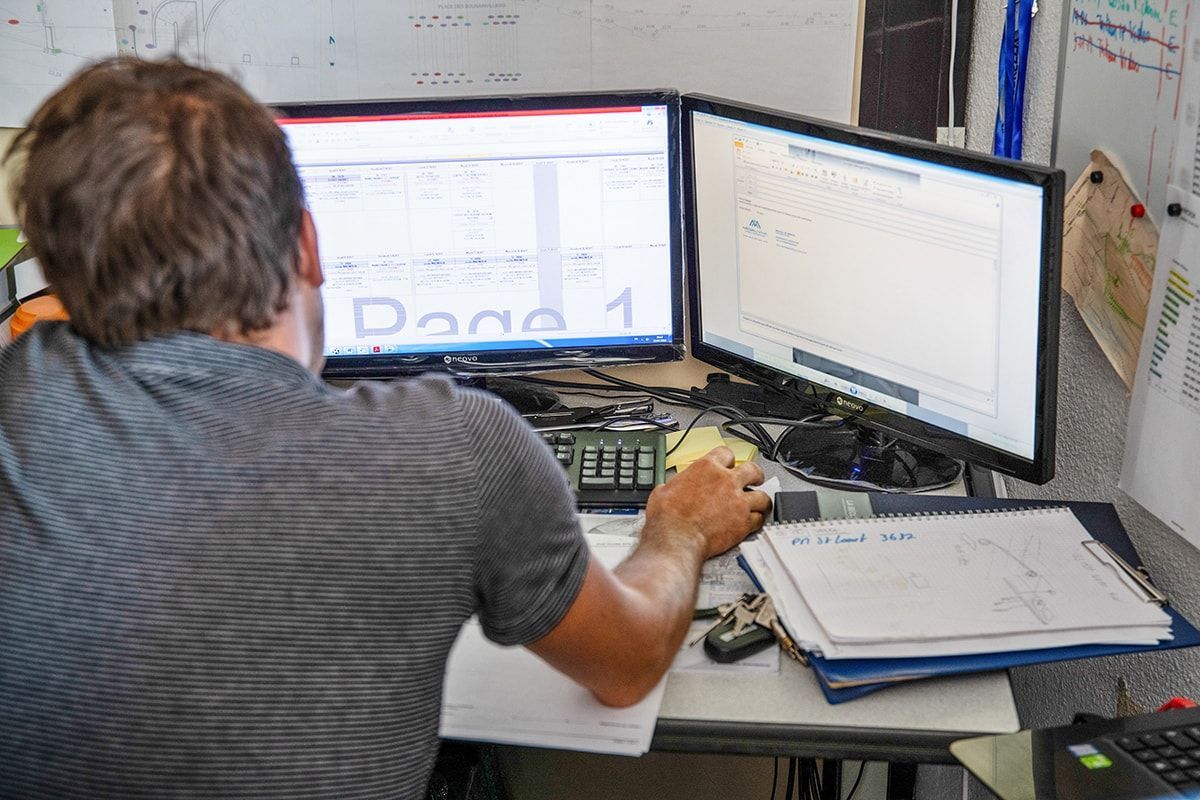 Personne travaillant à un bureau avec deux écrans, des documents et un clavier dans un environnement de bureau