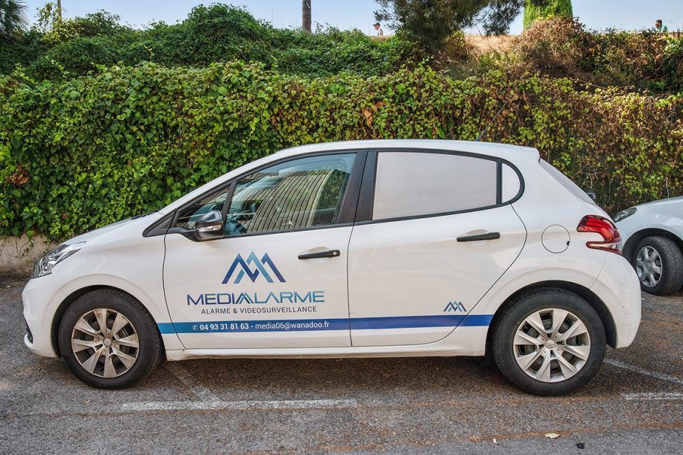 Voiture blanche à hayon avec le logo garée en bord de route, près de haies vertes.